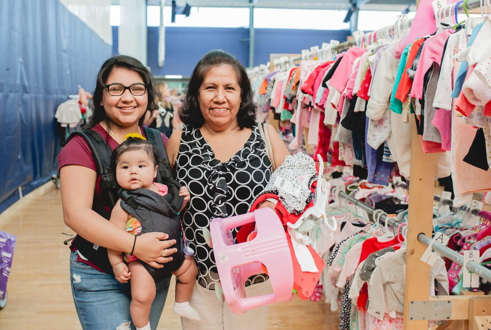 A mom with a large JBF shopping bag on her shoulder stands beside her husband who wears their toddler at a JBF sale.